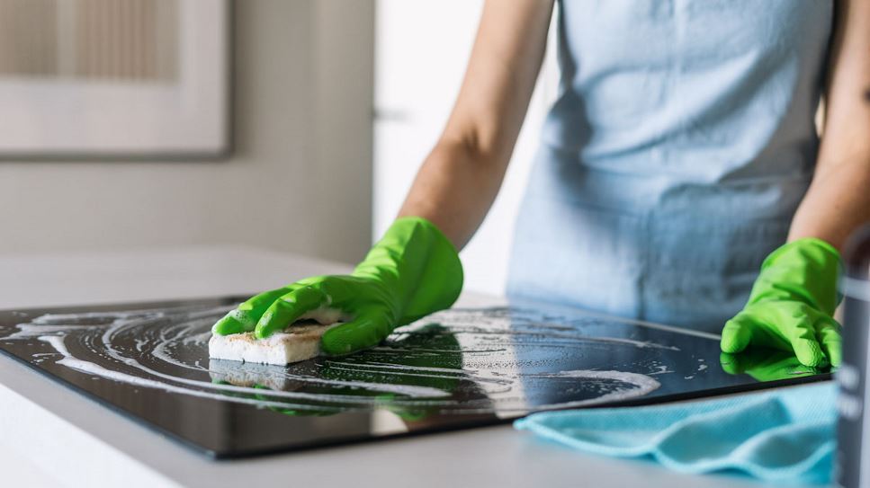 Individual in green cleaning gloves, cleaning induction cooktop with sponge