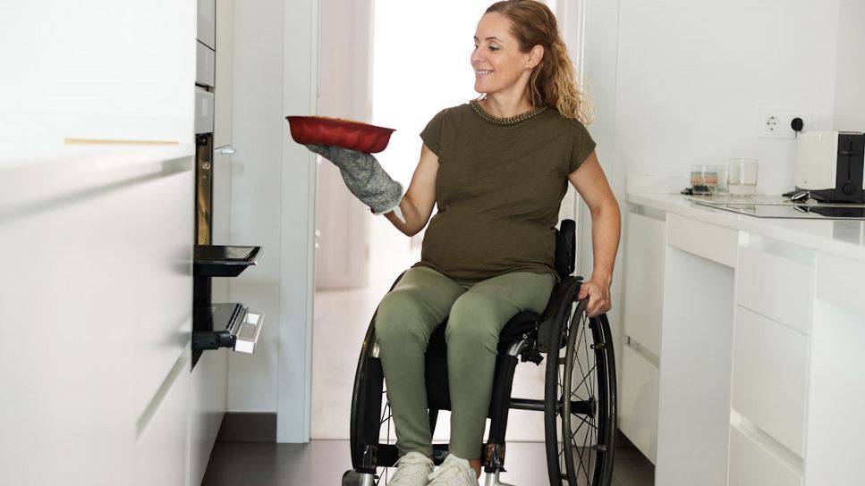 Pregnant woman in wheelchair, putting a cake into an adapted oven.
