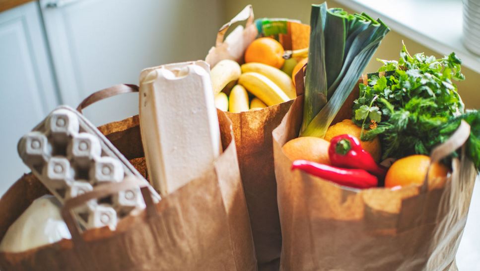 Three brown bags of groceries, sitting on a counter. They contain fresh produce, including chili peppers, leeks, bananas and citrus, as well as a carton of eggs.